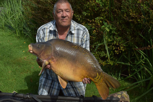 Photo of Ken holding a large carp on the riverside wearing a check shirt