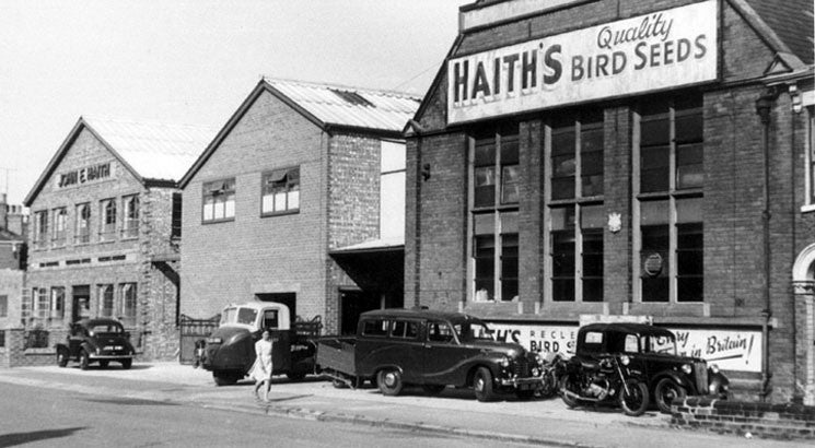 Image shows Haith's bird food factory in Cleethorpes during the 1950s with a Haith's Quality Bird Seeds sign suspended from the building and several vehicles awaiting coolection of bird seed. 